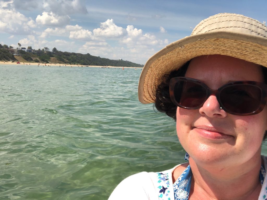 White woman with a hat and sunnies smiles in front of the water.